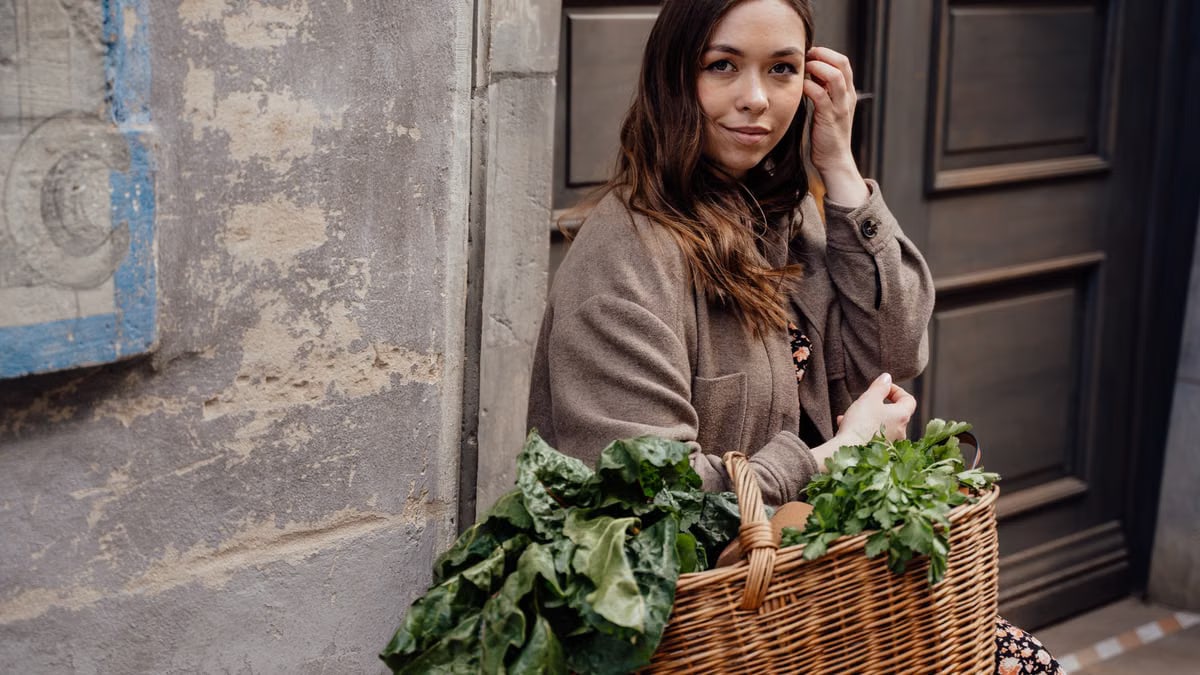 A woman smiling while holding a wicker basket filled with fresh green vegetables outdoors.